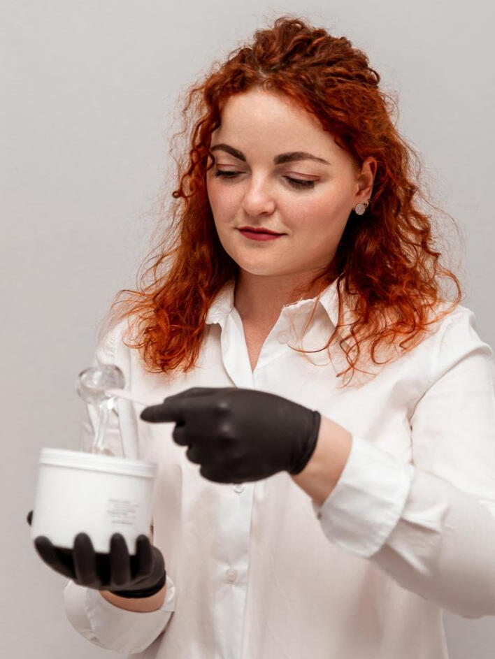 Jakość i bezpieczeństwo – nasz priorytet przy każdym zabiegu Kosmetolog o rudych włosach w białej koszuli i czarnych rękawiczkach demonstruje profesjonalną pastę do depilacji cukrowej